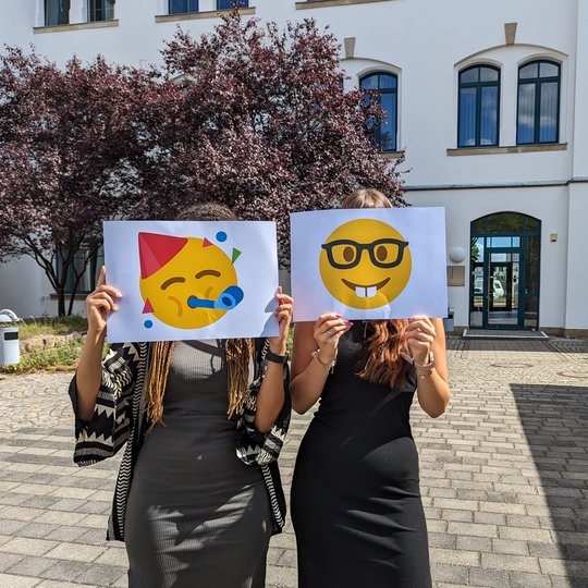 Zwei Mitarbeiterinnen vorm Bildungszentrum mit glücklichen Emoji-Schildern vorm Gesicht